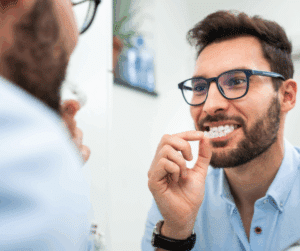 Smiling man with a beard and glasses looking in a mirror while holding up a clear teeth aligner to his mouth.