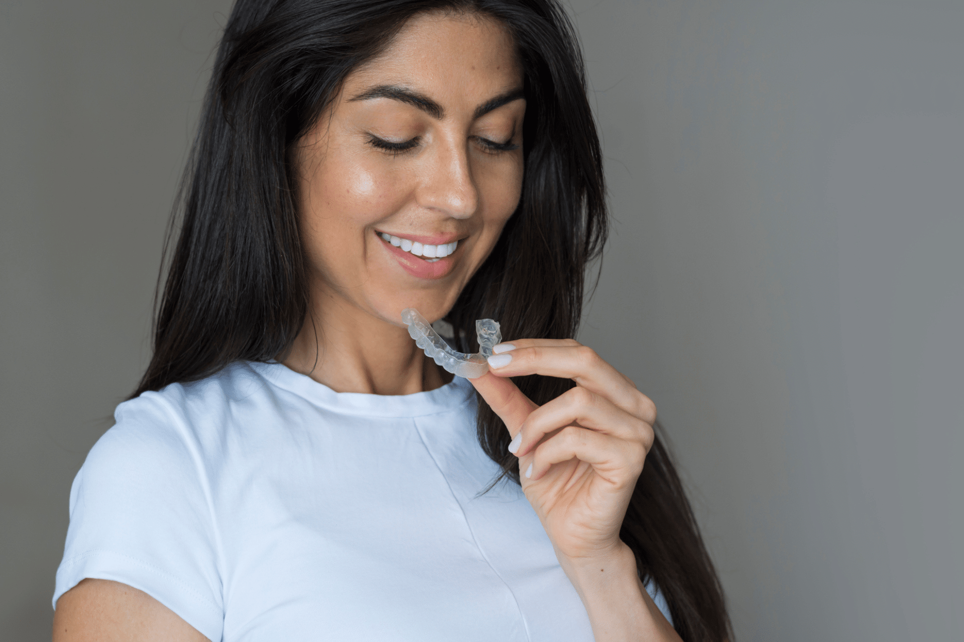 Smiling college-age woman holding a clear aligner against a gray background