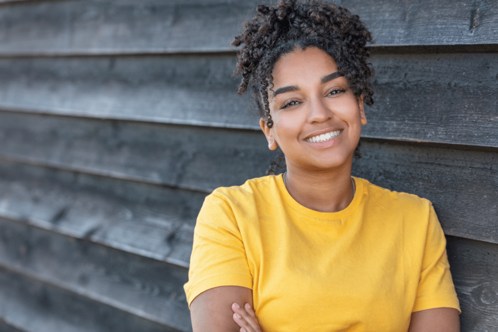 Smiling young woman in a yellow shirt leaning against a dark wooden wall