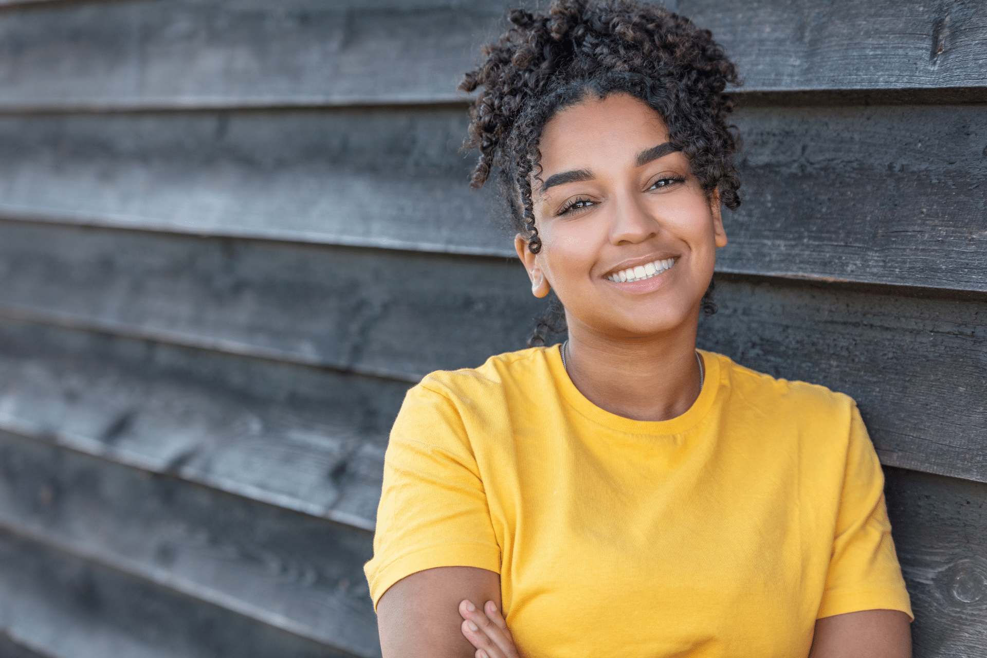 Smiling young woman in a yellow shirt leaning against a dark wooden wall